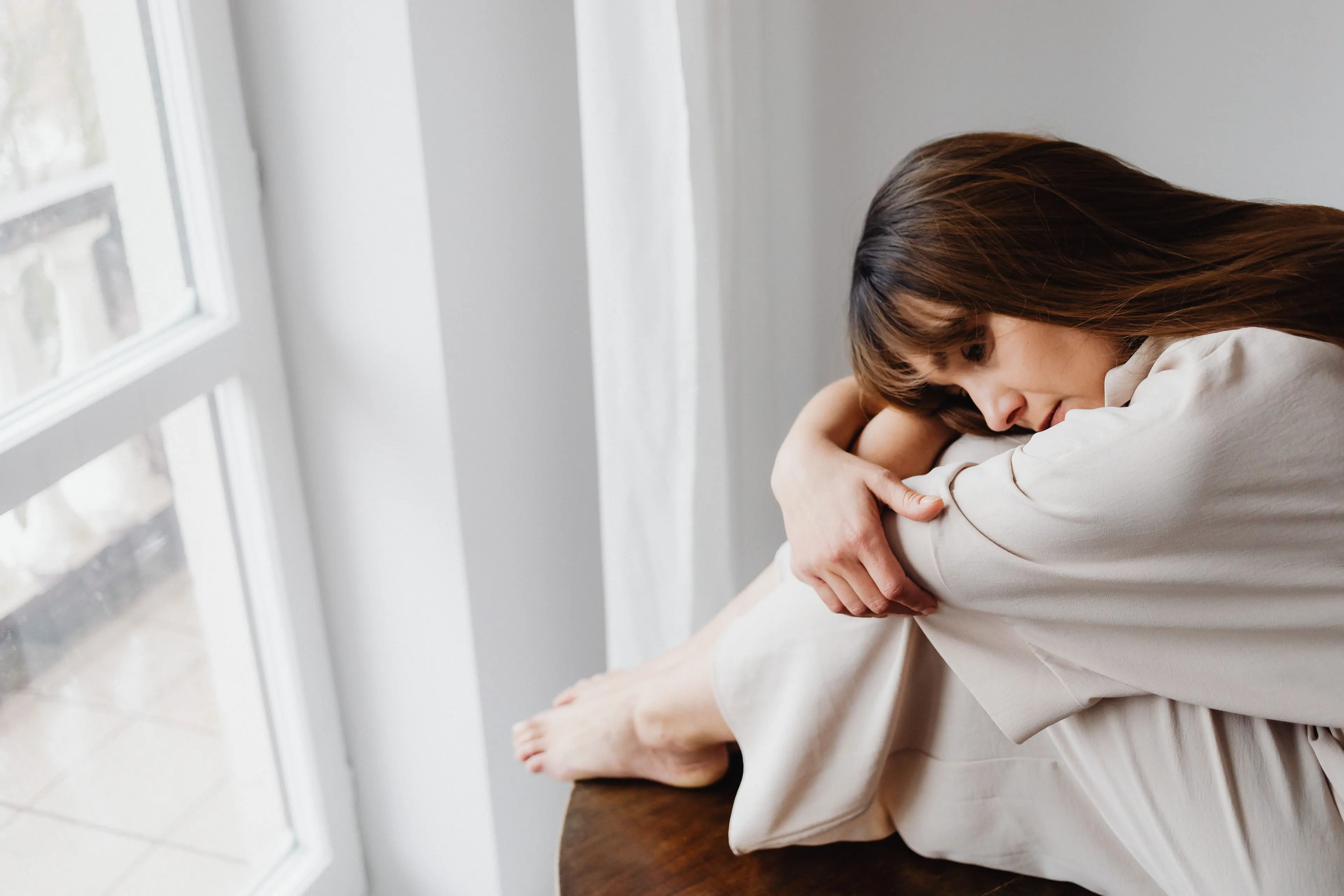 A woman sitting by a window, hugging her knees with her head resting on her arms — trauma therapy in Los Angeles
